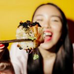Close-up of sushi being eaten with chopsticks by a woman with a bright yellow background.